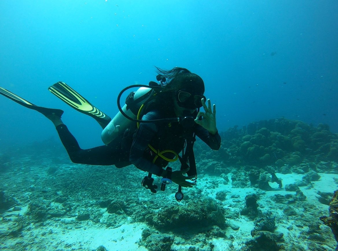 Sloka scuba diving over a coral reef, giving the OK hand sign